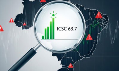 Imagem conceitual no estilo de infográfico. Uma grande lupa está posicionada sobre um mapa estilizado do Brasil. Dentro da lupa, a cena é clara e otimista, com um gráfico de barras verde subindo e o texto "ICSC 63,7". Fora da lupa, o mapa do Brasil tem um filtro mais escuro e acinzentado, com linhas de gráficos voláteis e símbolos de alerta, representando a incerteza econômica. A imagem simboliza a confiança específica do setor de consórcios em contraste com a cautela econômica geral. Alta resolução, proporção 16:9.