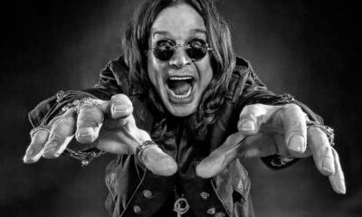 Um jovem Ozzy Osbourne, no final dos anos 60, em preto e branco, em uma encruzilhada de uma rua industrial de Birmingham. De um lado, uma fábrica sombria; do outro, um palco com uma luz brilhante. Ele está olhando para o palco com uma expressão de determinação. O estilo é de uma foto de filme granulado, dramático, alta resolução, proporção 16:9.
