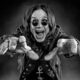 Um jovem Ozzy Osbourne, no final dos anos 60, em preto e branco, em uma encruzilhada de uma rua industrial de Birmingham. De um lado, uma fábrica sombria; do outro, um palco com uma luz brilhante. Ele está olhando para o palco com uma expressão de determinação. O estilo é de uma foto de filme granulado, dramático, alta resolução, proporção 16:9.
