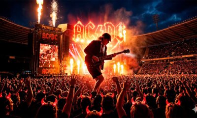 Uma fotografia épica e vibrante de um show de rock em um grande estádio lotado à noite. O foco principal é a silhueta enérgica de um guitarrista (remetendo à postura clássica de Angus Young com seu icônico uniforme escolar) no meio de um solo de guitarra explosivo, banhado por intensas luzes de palco vermelhas e amarelas (simbolizando a estética do AC/DC). Ao fundo, uma multidão eufórica com os braços erguidos e os característicos chifres luminosos vermelhos piscando na escuridão. A imagem deve transmitir eletricidade, paixão visceral e a imortalidade do rock. Estilo fotográfico hiper-realista e cinematográfico. Alta resolução, proporção 16:9 (820x468)