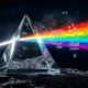Uma ilustração 3D conceitual e hiper-realista que homenageia o legado musical e a vida moderna. No centro, o icônico prisma triangular de vidro repousa sobre uma superfície texturizada, refratando um feixe de luz branca em um arco-íris brilhante e vibrante. Dentro do feixe de luz disperso, pequenas silhuetas e ícones holográficos flutuam sutilmente, representando a vida contemporânea: engrenagens de relógio (tempo), moedas flutuantes (ganância), rostos abstratos (ansiedade e mente frágil) e correntes se quebrando (liberdade e divisão social). O fundo é um espaço sideral profundo e escuro, contrastando com o brilho neon do prisma, transmitindo uma sensação de atemporalidade, introspecção e genialidade. Estilo de arte cinemático, nostálgico e profundo. Alta resolução, proporção 16:9 (820x468).