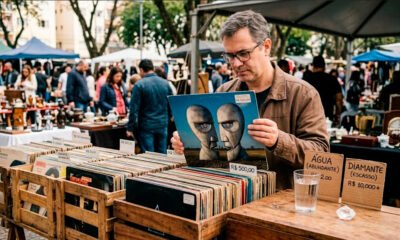 Fotografia com tom nostálgico de uma barraca em uma feira de antiguidades. Em foco principal, as mãos de uma pessoa seguram um disco de vinil antigo. Sobre a mesa rústica da barraca, compondo o cenário, repousam um copo de água e uma pedra brilhante semelhante a um diamante. A imagem cria uma metáfora visual para o paradoxo econômico do valor e da escassez.