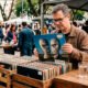 Fotografia com tom nostálgico de uma barraca em uma feira de antiguidades. Em foco principal, as mãos de uma pessoa seguram um disco de vinil antigo. Sobre a mesa rústica da barraca, compondo o cenário, repousam um copo de água e uma pedra brilhante semelhante a um diamante. A imagem cria uma metáfora visual para o paradoxo econômico do valor e da escassez.